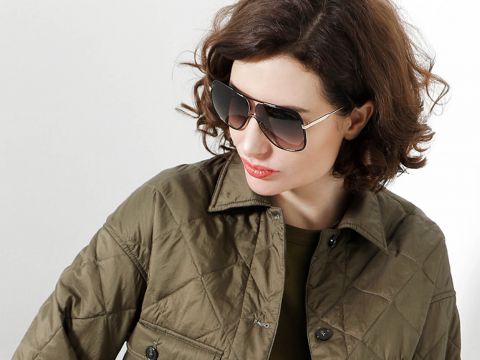 Eine Person mit welligem braunem Haar trägt eine große, dunkle Sonnenbrille und eine gesteppte olivgrüne Jacke. Sie blickt zur Seite vor einem schlichten Hintergrund.