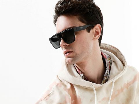 Mann mit großer schwarzer Sonnenbrille, Batik-Kapuzenpullover und gemustertem Hemd darunter, blickt nach links vor einem einfarbigen Hintergrund.