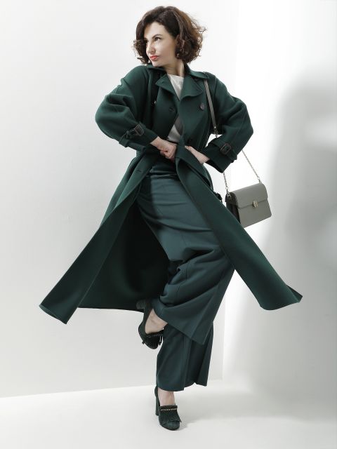 Eine Person steht selbstbewusst vor einem weißen Hintergrund, trägt einen stylischen dunkelgrünen Trenchcoat und eine dazu passende Hose. Sie trägt eine kleine grüne Umhängetasche und modische High Heels und nimmt eine dynamische Pose ein.