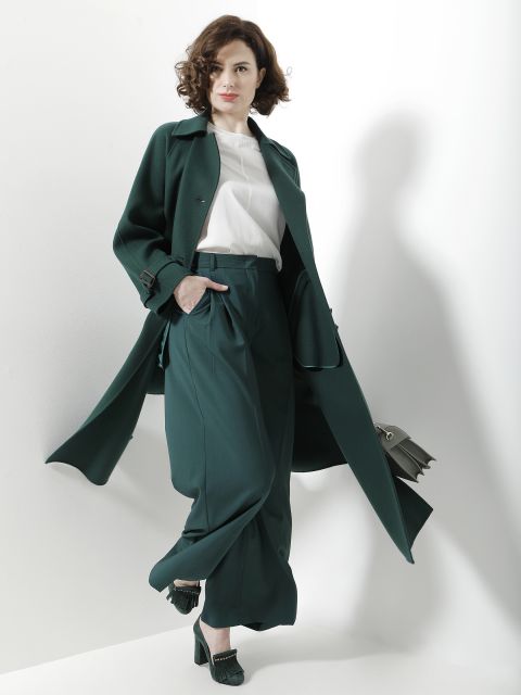 Eine Frau mit lockigem Haar posiert selbstbewusst vor einem weißen Hintergrund. Sie trägt einen stylischen dunkelgrünen Mantel und dazu passende weite Hosen, dazu eine weiße Bluse und dunkelgrüne Pumps. In der Hand hält sie eine hellgrüne Handtasche.