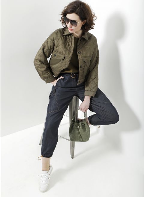 Eine Person mit Sonnenbrille, grüner Steppjacke und dunklen Jeans-Jogginghosen sitzt auf einem Hocker vor einem weißen Hintergrund. Sie hält eine grüne Handtasche in der Hand und trägt weiße Turnschuhe.