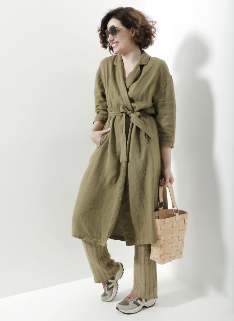 Eine Person mit kurzen braunen Haaren trägt eine Sonnenbrille und ein grünes Kleid mit Gürtel über gestreiften Hosen. Sie hält eine geflochtene Korbtasche und trägt Turnschuhe. Sie steht vor einem weißen Hintergrund.