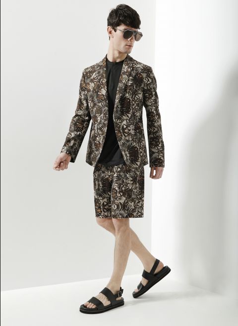 Ein Mann mit Sonnenbrille, gemustertem Blazer, passenden Shorts, schwarzem Hemd und Sandalen posiert vor einem weißen Hintergrund.