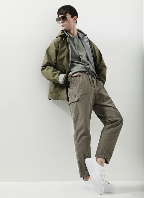 Eine Person mit dunkler Sonnenbrille und olivgrüner Jacke über einem grauen Kapuzenpullover posiert selbstbewusst vor einer weißen Wand. Sie trägt khakifarbene Cargohosen und weiße Turnschuhe und strahlt damit einen lässigen und stylischen Look aus.