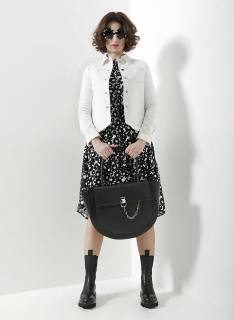 Eine Person steht selbstbewusst vor einer weißen Wand. Sie trägt eine weiße Jeansjacke über einem schwarzen Blumenkleid und hält eine große schwarze Handtasche in der Hand. Sie trägt eine schicke Sonnenbrille und Stiefeletten und schafft damit einen schicken, zeitgenössischen Look.