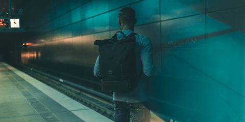 Eine Person mit einem Rucksack steht auf einem leeren U-Bahnsteig und blickt auf die Gleise. Die Station ist schwach mit blaugrünem Licht beleuchtet, was eine stimmungsvolle Atmosphäre schafft. Eine Uhr an der Decke zeigt die Zeit an, ein Zug ist nicht zu sehen.