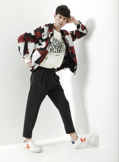 Eine Person posiert vor einer weißen Wand. Sie trägt eine rot-schwarz-weiß gemusterte Jacke, ein weißes Sweatshirt mit Text, eine schwarze Hose und weiße Turnschuhe mit roten und blauen Akzenten. Sie hat kurze dunkle Haare und nimmt eine selbstbewusste Pose ein.