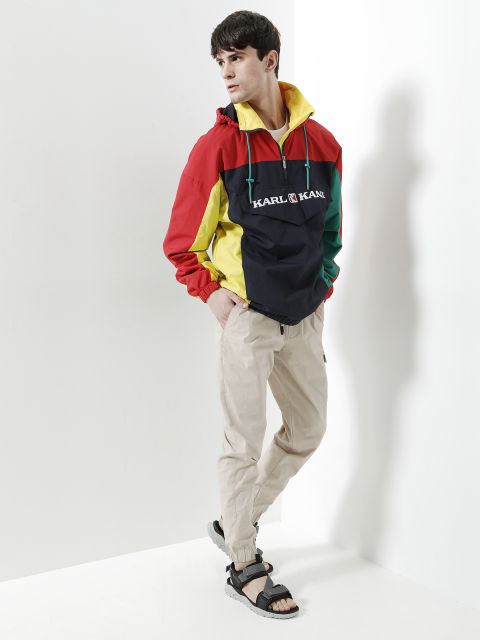 Eine Person steht lässig in einer Ecke und trägt eine bunte Windjacke mit roten, gelben, grünen und schwarzen Abschnitten und beige Hosen. Sie hat dunkle Haare und trägt schwarze Sandalen. Sie schaut zur Seite und hat eine Hand in der Tasche.