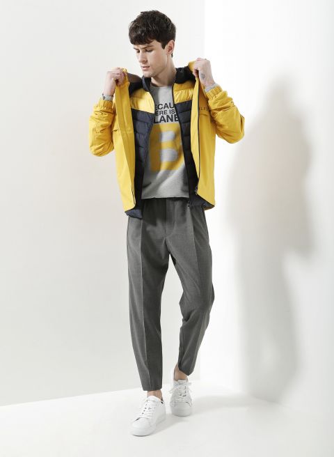 Eine Person steht vor einem weißen Hintergrund und trägt eine gelbe Jacke, ein graues T-Shirt mit großem Text, eine graue Hose und weiße Turnschuhe. Sie hält den Jackenkragen und blickt zur Seite.