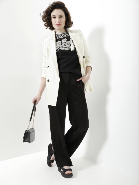 Eine Person mit lockigem Haar steht vor einer weißen Wand und trägt einen weißen Blazer über einem schwarzen bestickten Oberteil und eine schwarze Hose. Sie hält eine kleine Handtasche und trägt schwarze Sandalen. Sie posiert mit einer Hand in der Tasche.