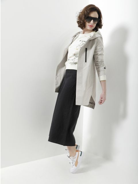 Eine Person mit lockigem Haar trägt eine Sonnenbrille, einen beigen Mantel, ein bedrucktes weißes Hemd, schwarze Hosen mit weitem Bein und weiße Turnschuhe. Sie posieren stilvoll vor einer weißen Wand.