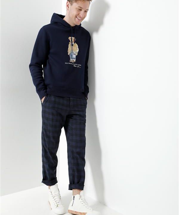 Eine Person steht lächelnd vor einer weißen Wand. Sie trägt einen dunkelblauen Kapuzenpullover mit Bärenmotiv, karierte Hosen und weiße Turnschuhe. Sie hat die Hände in den Taschen und ihr blondes Haar ist lässig gestylt.