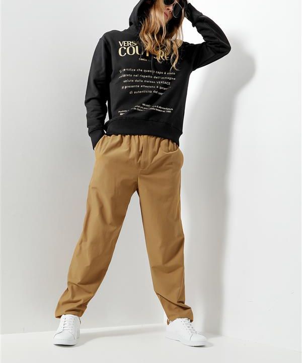 Eine Person posiert in einem stylischen Modeoutfit. Sie trägt einen schwarzen Kapuzenpullover mit Text, beige Hosen und weiße Turnschuhe. Sie hat wallendes Haar und trägt eine Sonnenbrille. Sie steht vor einem schlichten weißen Hintergrund.