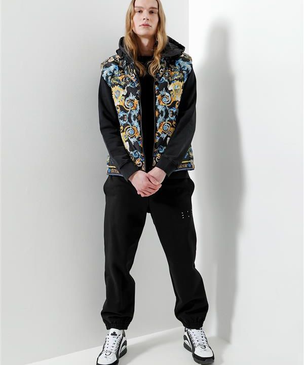 Eine Person mit langen Haaren steht vor einer weißen Wand. Sie trägt einen schwarzen Kapuzenpullover, eine bunt gemusterte Weste, eine schwarze Hose und weiße Turnschuhe. Die Hände sind vor dem Körper gefaltet.