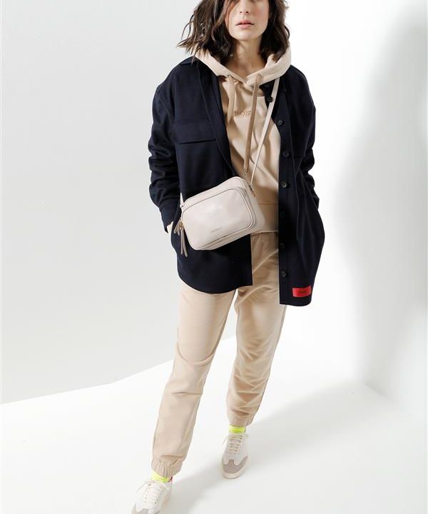 Eine Person in einem beigen Kapuzenpullover und einer beigen Hose mit neonfarbenen Schuhen steht vor einem weißen Hintergrund. Sie trägt eine schwarze Jacke und eine beige Umhängetasche.