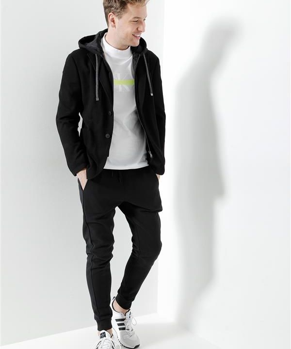 Eine Person in lässiger Kleidung steht vor einem weißen Hintergrund. Sie trägt einen schwarzen Kapuzenpullover, ein weißes T-Shirt, schwarze Jogginghosen und weiße Turnschuhe. Die Person schaut zur Seite und lächelt, die Hände in den Taschen.