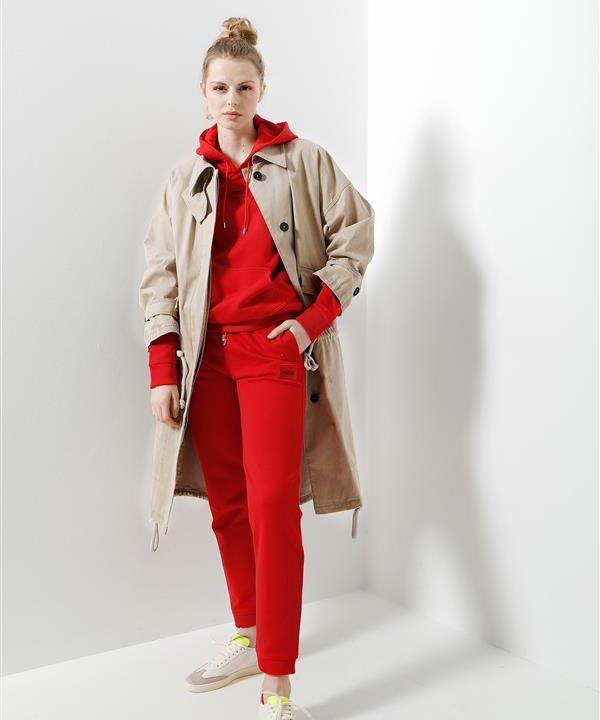 Eine Person steht in einer Ecke. Sie trägt einen langen beigefarbenen Trenchcoat über einem roten Kapuzenpullover und einer roten Hose. Sie trägt außerdem weiße Turnschuhe mit gelben Details. Der Hintergrund ist eine einfache weiße Wand.