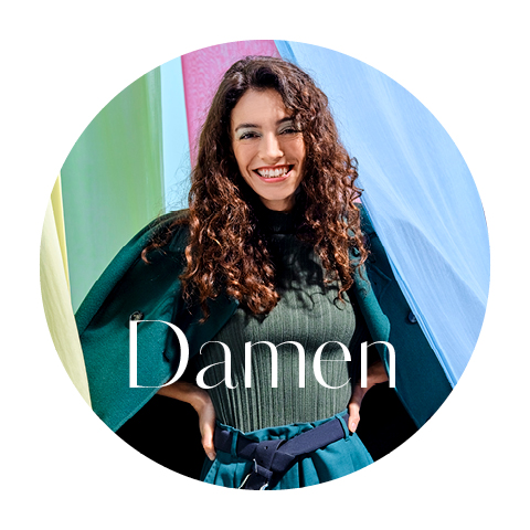 Eine Frau mit lockigem Haar steht lächelnd vor bunten Stoffbahnen. Sie trägt ein dunkelgrünes Outfit und einen Gürtel. Das Wort „Damen“ ist über das Bild gelegt.