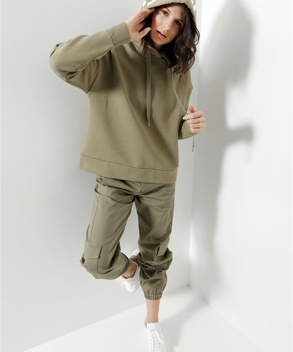 Person mit einem khakifarbenen Kapuzenpullover mit hochgezogener Kapuze, dazu passenden Cargo-Jogginghosen und weißen Turnschuhen steht in einem minimalistischen Raum mit weißen Wänden. Sie blickt direkt in die Kamera und hat eine Hand auf der Kapuze.