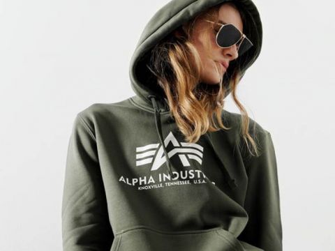 Eine Person mit langen Haaren, die eine Pilotenbrille und einen olivgrünen Kapuzenpullover mit „Alpha Industries“ und einem Logo auf der Vorderseite trägt. Die Kapuze ist hochgezogen und sie blickt nach unten und zur Seite. Der Hintergrund ist schlicht weiß.