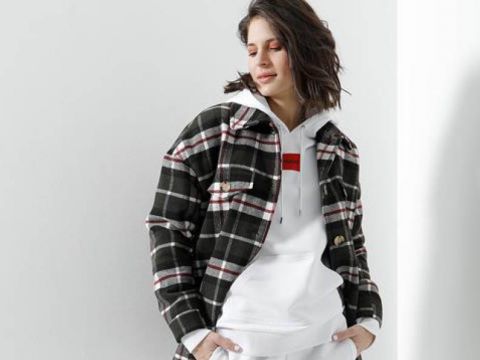 Eine Person, die eine karierte Jacke über einem weißen Kapuzenpullover trägt, steht vor einem weißen Hintergrund. Die Jacke weist ein schwarz-weißes Karomuster mit roten Akzenten auf. Die Person hat schulterlanges dunkles Haar und scheint nach unten zu schauen.