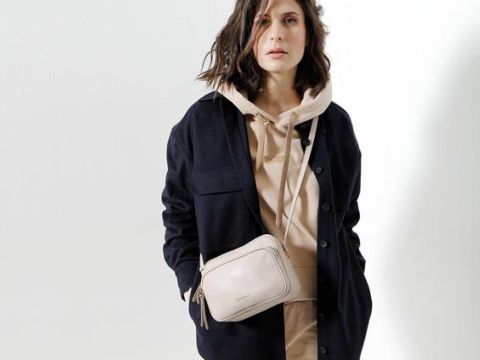 Eine Person mit schulterlangem Haar trägt einen beigen Kapuzenpullover und eine schwarze Jacke, dazu eine beige Umhängetasche. Sie stehen vor einem schlichten weißen Hintergrund.