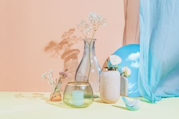 Eine Stilllebenkomposition mit Glasvasen mit zarten Blumen, einem runden Spiegel, der einen blauen Himmel mit Wolken reflektiert, einem hellblauen Vorhang und einem kleinen vogelförmigen Ornament vor einem pastellfarbenen pfirsich- und gelben Hintergrund.