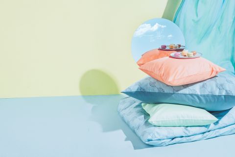 Ein Stapel pastellfarbener Kissen auf einer blauen Oberfläche mit einer gelben Wand dahinter. Ein runder Spiegel reflektiert den Himmel und auf dem obersten Kissen liegt ein Teller mit bunten Macarons. Im Hintergrund hängt ein fließender, transparenter Stoff.