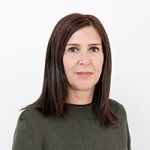 Eine Frau mit schulterlangem braunem Haar und einem dunkelgrünen Pullover steht vor einem schlichten weißen Hintergrund. Sie blickt mit neutralem Gesichtsausdruck in die Kamera.