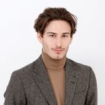 Eine Person mit braunem Haar und Schnurrbart trägt einen grauen Blazer über einem beigen Rollkragenpullover. Sie lächelt leicht und blickt vor einem schlichten weißen Hintergrund in die Kamera.