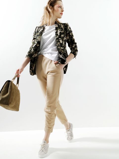 Eine Frau posiert stilvoll in beigen Jogginghosen, einem weißen Hemd und einer Camouflage-Jacke. Sie hält eine hellbraune Handtasche und trägt weiße Turnschuhe. Ihr Haar weht im Wind vor einem schlichten Hintergrund.