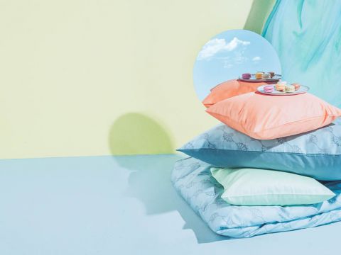 Auf einem Stapel pastellfarbener Kissen liegt ein Spiegel, der einen blauen Himmel und Wolken reflektiert. Auf dem Spiegel liegen Macarons auf einem Teller. Der Hintergrund ist in sanftem Gelb und hellem Türkis gehalten und schafft eine heitere und verträumte Atmosphäre.
