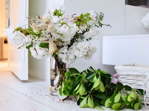Ein Strauß weißer Blumen mit Grünpflanzen steht in einer durchsichtigen Glasvase auf einem hellen Holzboden. Daneben sind weitere grüne Blätter und ein weißer Weidenkorb zu sehen, die eine ruhige, minimalistische Innenatmosphäre schaffen.