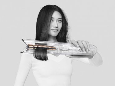 Eine Frau mit langen, glatten schwarzen Haaren hält einen transparenten Haarglätter. Sie trägt einen weißen Pullover und steht vor einem schlichten Hintergrund. Ihr Gesichtsausdruck ist ruhig und selbstbewusst.