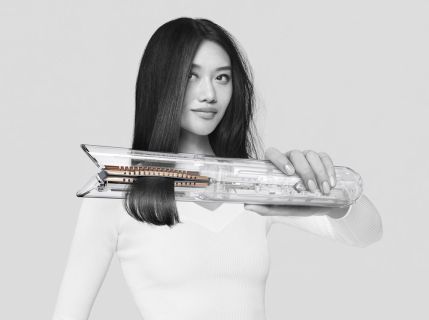 Eine Frau mit langen, glatten schwarzen Haaren hält einen transparenten Haarglätter. Sie trägt einen weißen Pullover und steht vor einem schlichten Hintergrund. Ihr Gesichtsausdruck ist ruhig und selbstbewusst.