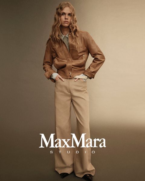 Eine Person mit langen Haaren trägt eine braune Lederjacke über einem hellen Hemd und dazu beigefarbene Hosen mit weitem Bein. Sie stehen vor einem beigefarbenen Hintergrund mit Farbverlauf. Unten ist der Text „MaxMara Studio“ zu sehen.