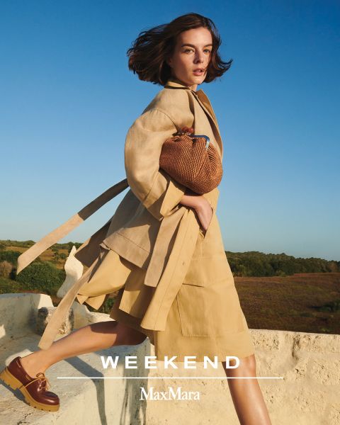 Eine Person in einem beigen Mantel und einem dazu passenden Rock steht auf einem Felsvorsprung vor einem klaren blauen Himmel und hält eine strukturierte braune Tasche in der Hand. Unten steht der Text „WEEKEND MaxMara“.