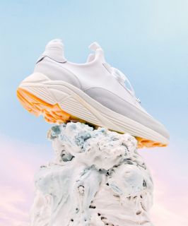 Ein stylischer Sneaker in Weiß- und Hellgrau-Design mit dicker Sohle und orangefarbenen Akzenten steht auf einem strukturierten, abstrakten weißen Sockel vor einem Himmelshintergrund mit Farbverlauf in Blau und Rosa.
