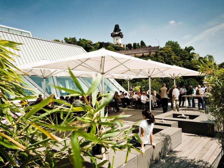 Menschen genießen einen sonnigen Tag in einem Straßencafé mit großen Sonnenschirmen. Im Hintergrund thront ein historischer Glockenturm auf einem grünen Hügel. Üppiges Laub und moderne Architektur umgeben den Sitzbereich.