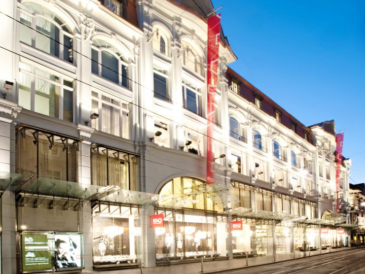 Ein großes, beleuchtetes Kaufhaus mit einer prachtvollen Fassade, vielen gewölbten Fenstern und roten Bannern. Die Straße davor ist ruhig und der Himmel ist klar und blau, was auf die Dämmerung hindeutet.