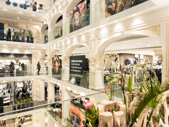 Ein elegantes mehrstöckiges Einkaufszentrum mit klassischen architektonischen Details. Käufer können in Geschäften mit großen Glasfenstern nach Kleidung stöbern. Um das offene Atrium herum sind dekorative Pflanzen und Blumen aufgestellt.