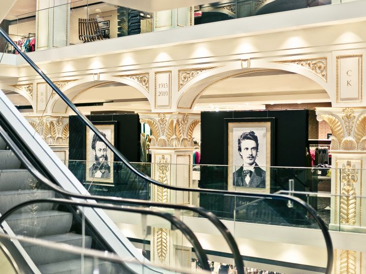 Innenansicht eines kunstvoll verzierten Kaufhauses mit Rolltreppen und zwei gerahmten Porträts eines Mannes, die im oberen Stockwerk ausgestellt sind. Der Raum zeichnet sich durch dekorative Säulen und elegante Beleuchtung aus.