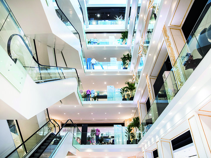 Innenansicht eines modernen mehrstöckigen Einkaufszentrums mit heller Beleuchtung, Glasgeländern, Rolltreppen und Grünflächen auf jeder Etage. Das offene Design präsentiert mehrere Etagen und Ladenfronten in einer geräumigen Umgebung.