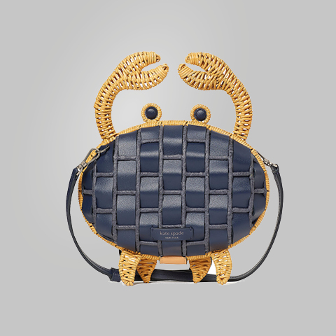 Eine skurrile Handtasche, die einer Krabbe ähnelt und eine gewebte Textur in Marineblau- und Goldtönen aufweist. Die Tasche hat oben zwei krabbenähnliche Scheren und unten sechs Beine und ist mit einem Schultergurt versehen.