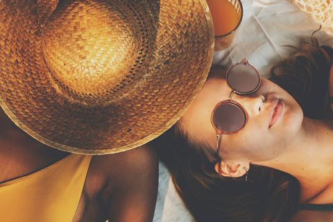Zwei Menschen entspannen auf einer Stranddecke. Einer trägt einen großen Strohhut, der sein Gesicht verdeckt, der andere trägt eine Sonnenbrille und liegt daneben. Beide tragen Badebekleidung und haben ein Glas Orangensaft in der Nähe, was für eine sommerliche und entspannte Stimmung sorgt.