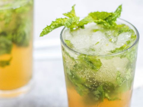 Ein erfrischender Mojito in einem hohen Glas, garniert mit frischen Minzblättern. Das Getränk ist leicht golden mit sichtbaren Minzblättern und Eiswürfeln, was ihm ein frostiges, appetitanregendes Aussehen verleiht. Ein weiterer Mojito ist im Hintergrund leicht verschwommen.