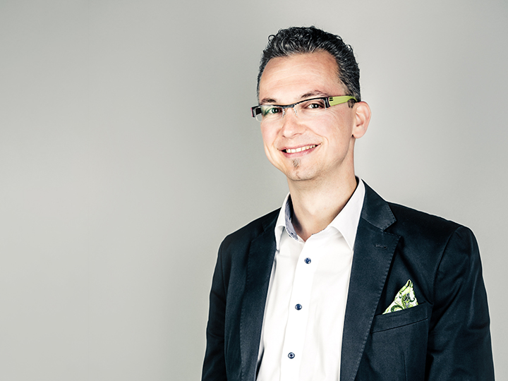 Ein Mann mit kurzen dunklen Haaren und Brille lächelt in die Kamera. Er trägt einen dunklen Blazer über einem weißen Hemd mit einem gemusterten Einstecktuch. Der Hintergrund ist schlicht hellgrau.