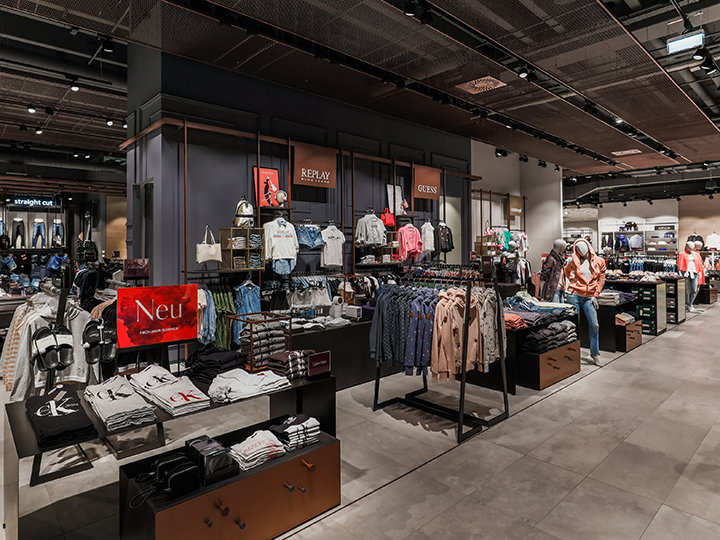 Ein modernes Bekleidungsgeschäft mit verschiedenen Abteilungen, in denen Hemden, Jacken und Accessoires ordentlich präsentiert werden. Das Geschäft zeichnet sich durch helle Beleuchtung, übersichtliche Auslagen und Markenbeschilderung aus und schafft so eine elegante und einladende Einkaufsumgebung.