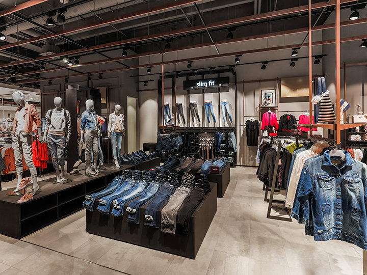 Innenansicht eines Bekleidungsgeschäfts mit Schaufensterpuppen in Jeans-Outfits. Auf Schautischen werden gefaltete Jeans präsentiert und auf Regalen stehen verschiedene Kleidungsstücke. Das Geschäft ist modern gestaltet, mit Industriebeleuchtung und einem Schild mit der Aufschrift „Slim Fit“.