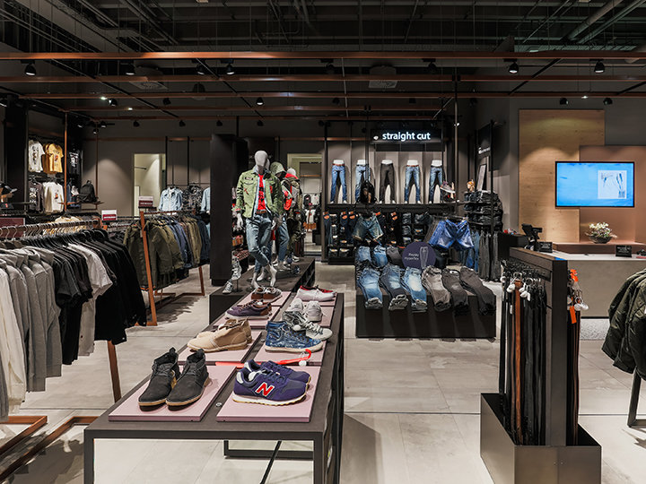Ein modernes Interieur eines Bekleidungsgeschäfts mit verschiedenen Kleidungsstücken, darunter Hemden, Jacken und Jeans. Schaufensterpuppen präsentieren Outfits, Schuhe sind auf Tischen angeordnet und im Hintergrund ist ein digitaler Bildschirm zu sehen. Auf dem Schild steht „gerader Schnitt“.